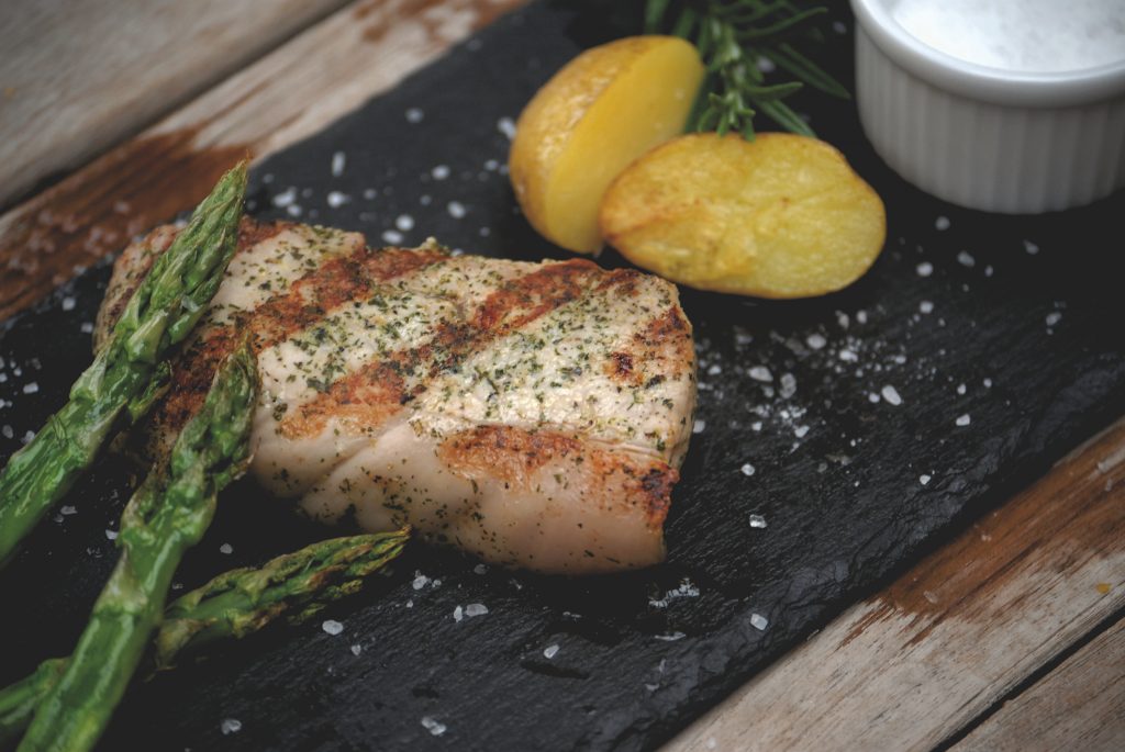 Foto eines zubereiteten marinierten Kalbsteak der Gassner GmbH, präsentiert auf einer schwarzen Schieferplatte, angerichtet mit gegrillten grünen Spargel und Bratkartoffeln