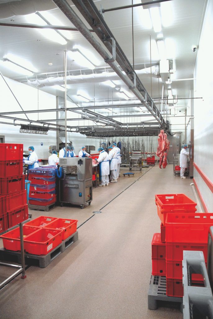 Aufnahme des Betriebes der Gassner GmbH. Im Vordergrund befinden sich rote Plastikkisten, im linken Hintergrund des Bildes sieht man Fleischer bei der Arbeit am Fleischtisch, im rechten Bildhintergrund sieht man Kalb Karkassen hängend.